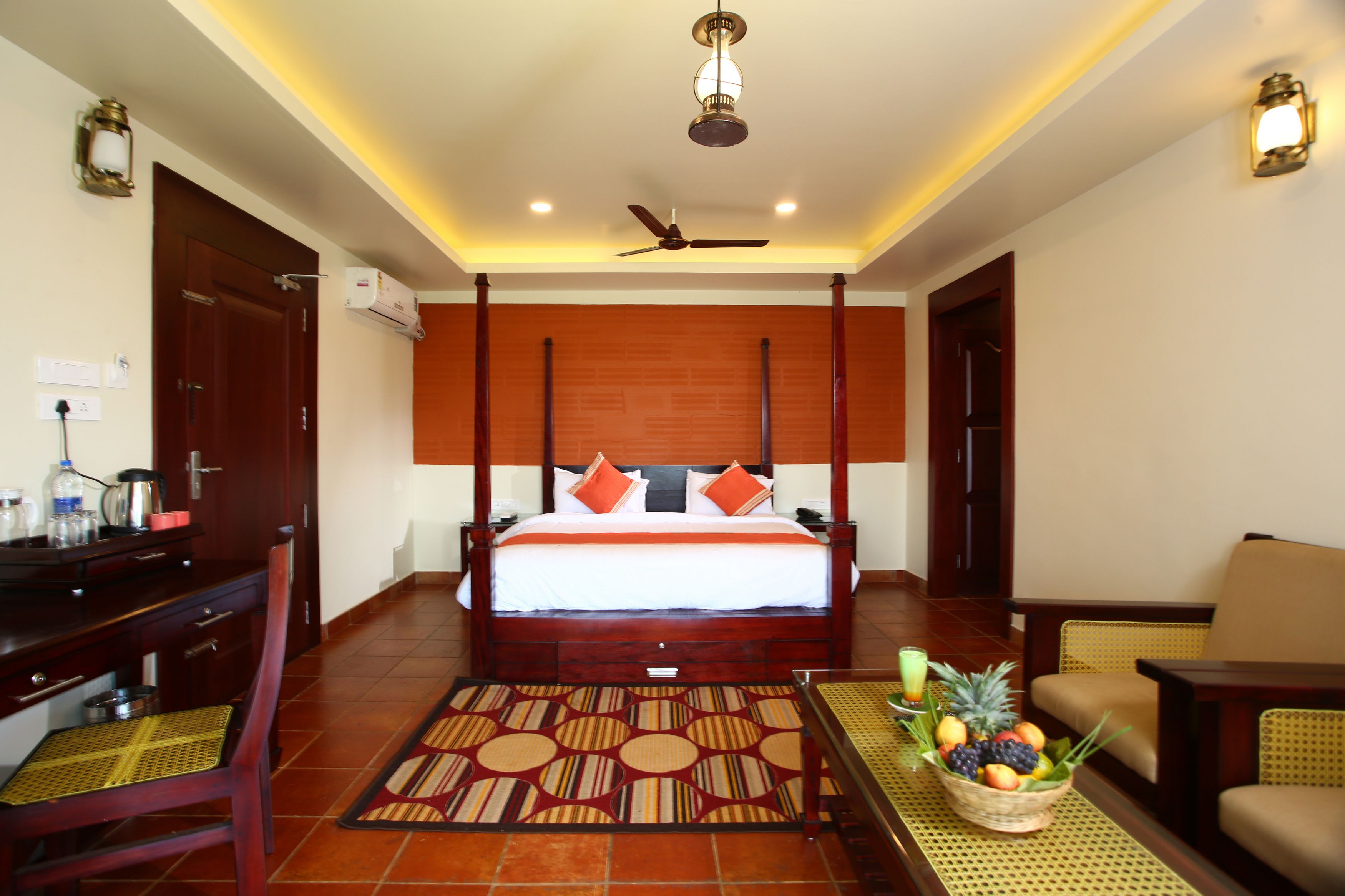 A hotel room with a four-poster bed, a desk with a chair, a dining table with a fruit basket, and a ceiling fan.