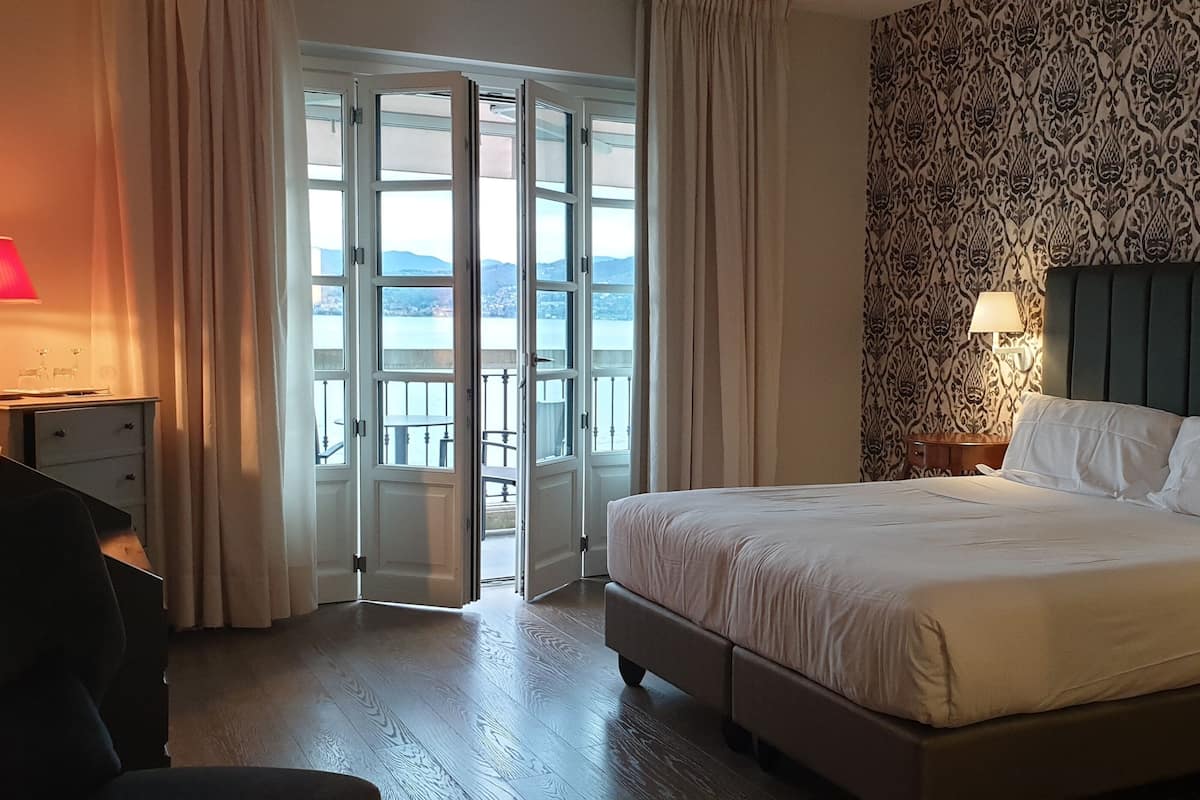 A hotel room with a bed, a desk, a chair, a lamp, and a view through a door.