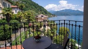 A balcony with a round table and a bowl of flowers, overlooking a lake with boats and houses in the background. - Park Hotel Italia (Cannero Riviera)