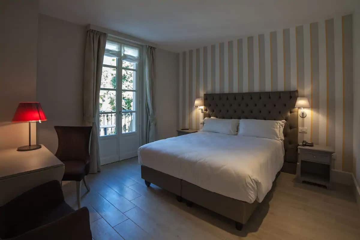 A hotel room with a large bed, two bedside tables with lamps, a chair, a desk, and a window with curtains.