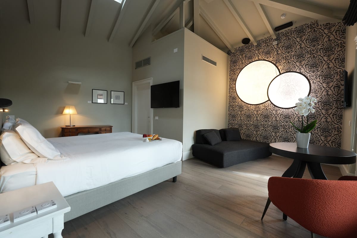 A modern bedroom with a large bed, a flat-screen TV, a dining table with a potted plant, and a patterned wall.