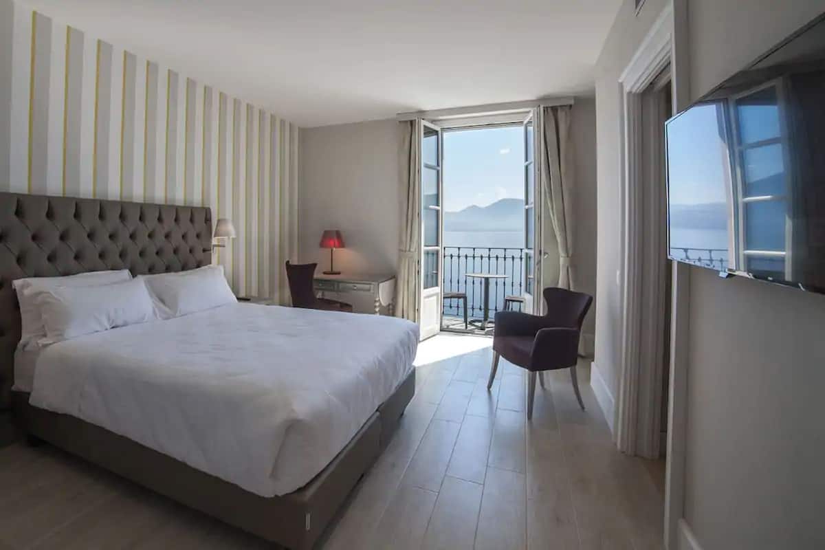 A hotel room with a large bed, a desk with a lamp, a chair, and a balcony with a view.