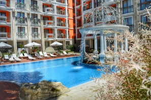 Area piscina di un hotel con diverse poltrone prendisole, una fontana al centro e balconi con ringhiere.