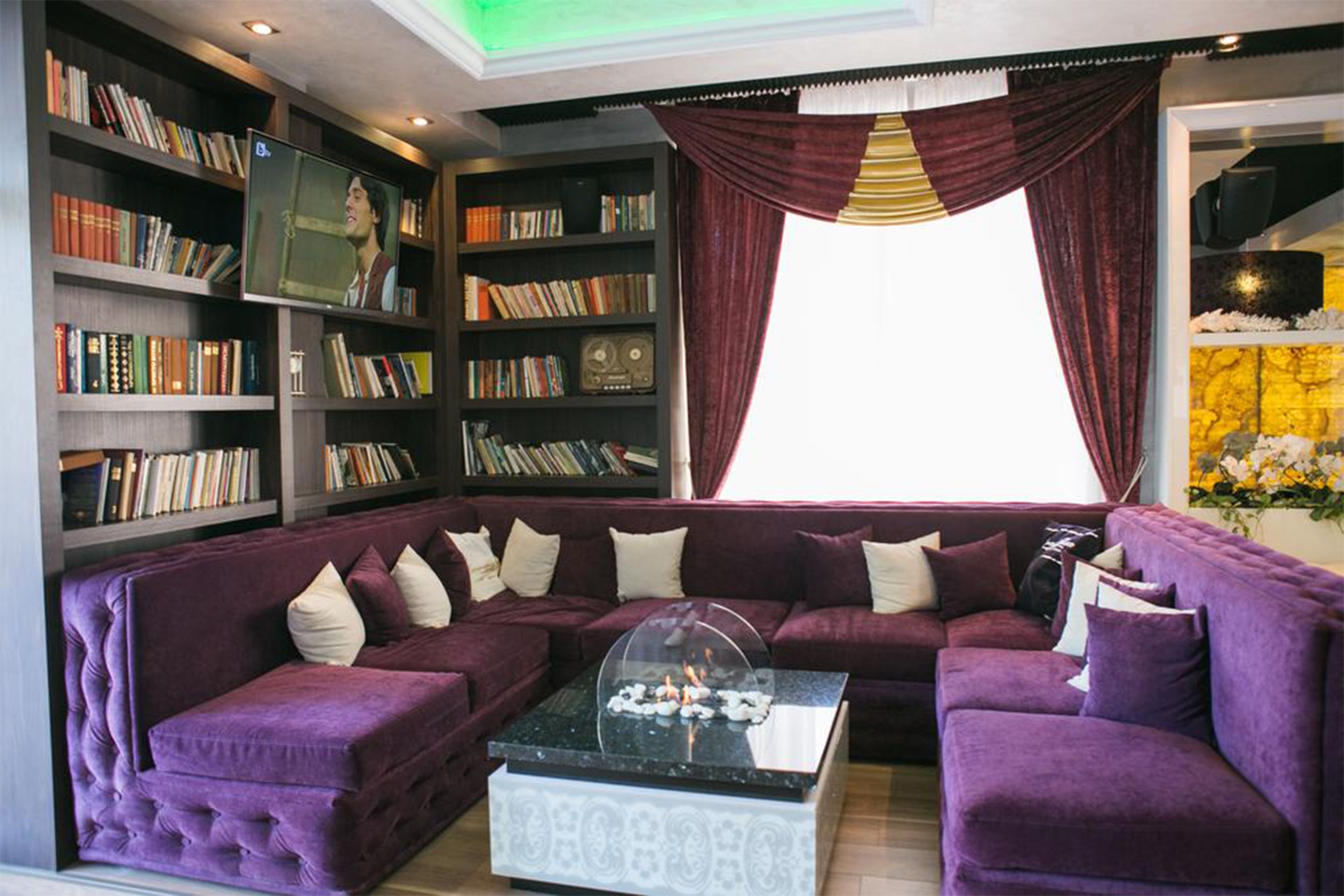 Ein Wohnzimmer mit einem violetten Sofagarnitur, einem Couchtisch mit Glaskuppel und Bücherregalen voller Bücher und einem Vintage-Radio.