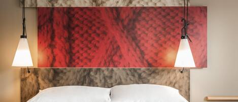 Habitación de hotel con cama, almohadas blancas, mesita de noche con teléfono y un cuadro de textura roja y beige en la pared.