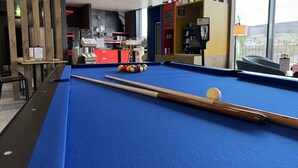 Ein Billardtisch mit Billardkugeln und Spielstöcken, eine rote Kommode und ein Fenster mit Blick auf eine gebirgige Landschaft.