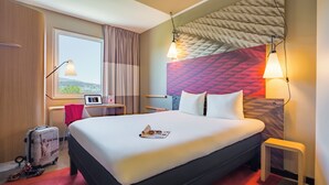 Een hotelkamer met een groot bed, een bureau met een computer, een koffer en een raam met gordijnen.