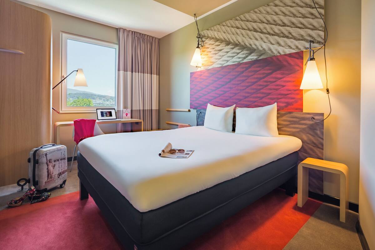 Une chambre d’hôtel avec un grand lit, un bureau avec un ordinateur, une valise et une fenêtre avec des rideaux.