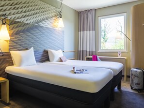 A hotel room with two beds, a window with curtains, a lamp, and a suitcase.