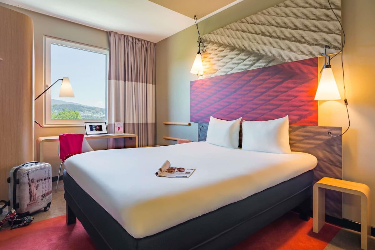 Ein Hotelzimmer mit einem großen Bett, einem Schreibtisch, einem Stuhl, einem Koffer und einem Vorhangfenster.