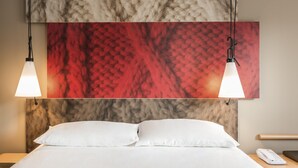A hotel room with a bed, white pillows, a nightstand with a phone, and a wall with a red and beige textured artwork.