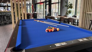 Ein Billardtisch mit blauer Spielvliesdecke, umgeben von Holzpaneelen und einer Sitzecke mit Tischen und Stühlen.