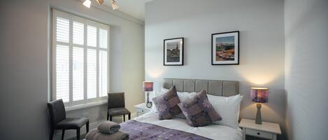A neatly arranged bedroom with a large bed, two bedside tables, a chair, and a window with blinds.