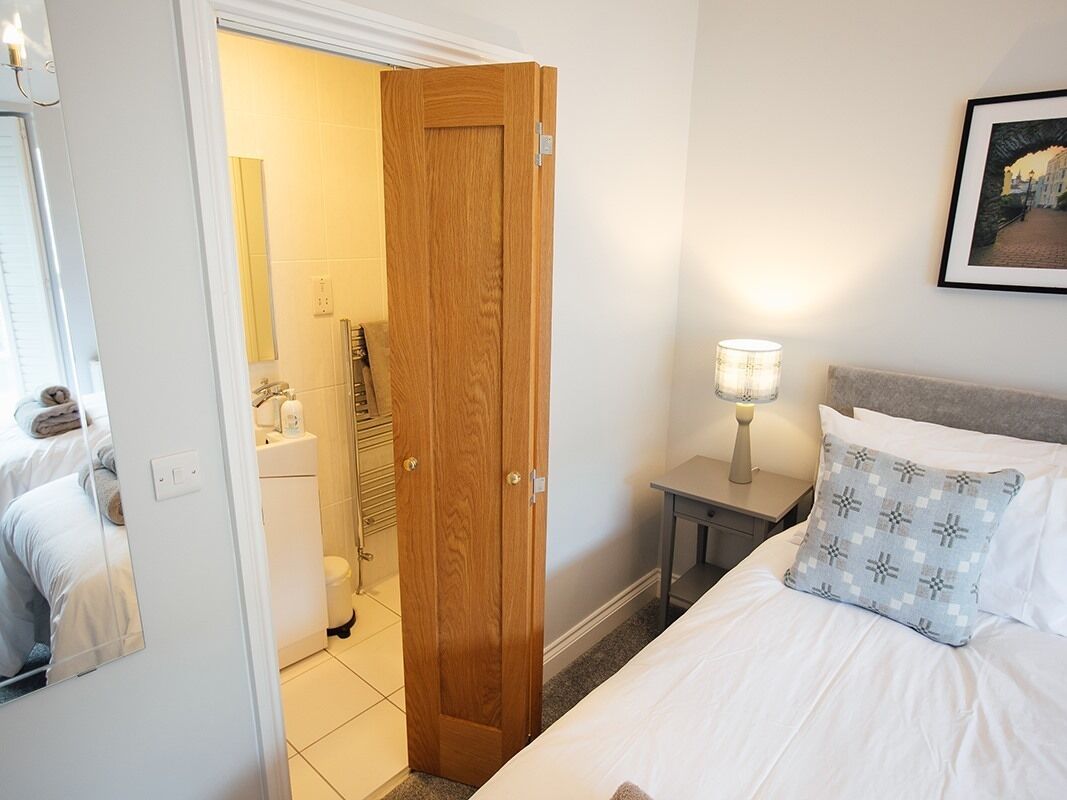 A hotel room with a wooden door, a bed with white bedding, a nightstand with a lamp, and a framed picture on the wall.