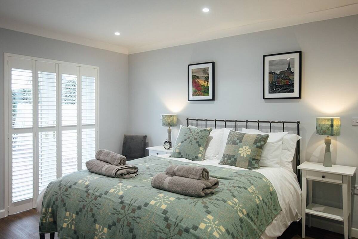 Un lit bien fait, recouvert d’une couvre-lit à motifs verts, avec deux oreillers et des serviettes pliées. Deux tableaux encadrés sont accrochés au mur, il y a une table de chevet avec une lampe, et une fenêtre avec des stores.