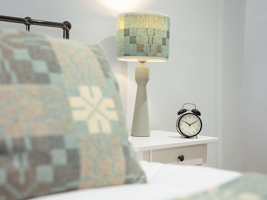 A neatly made bed with a patterned pillow, a white bedside table with a lamp and an alarm clock.