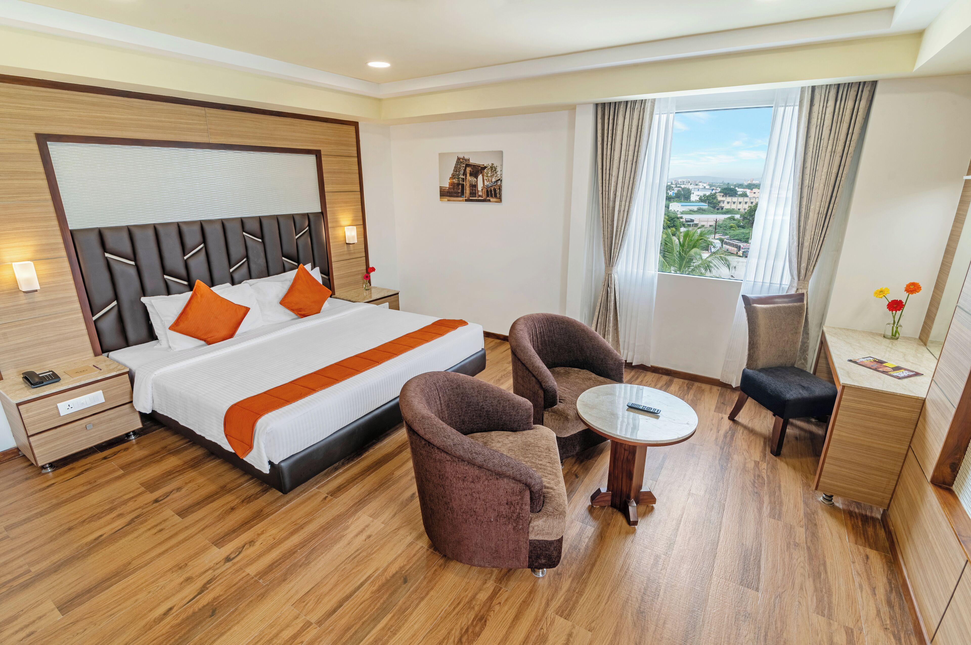 A hotel room with a large bed, two armchairs, a small table, a nightstand, and a view of the city through the window.