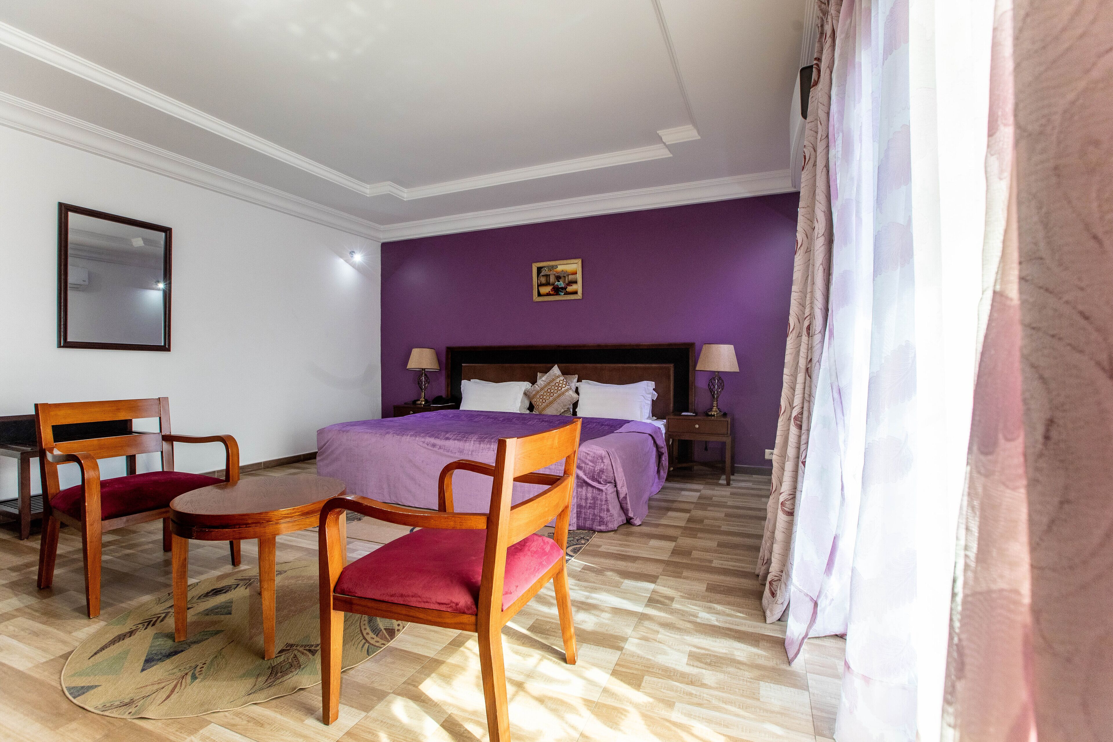 A hotel room with a bed, two wooden chairs, a small table, a nightstand, and a purple wall.