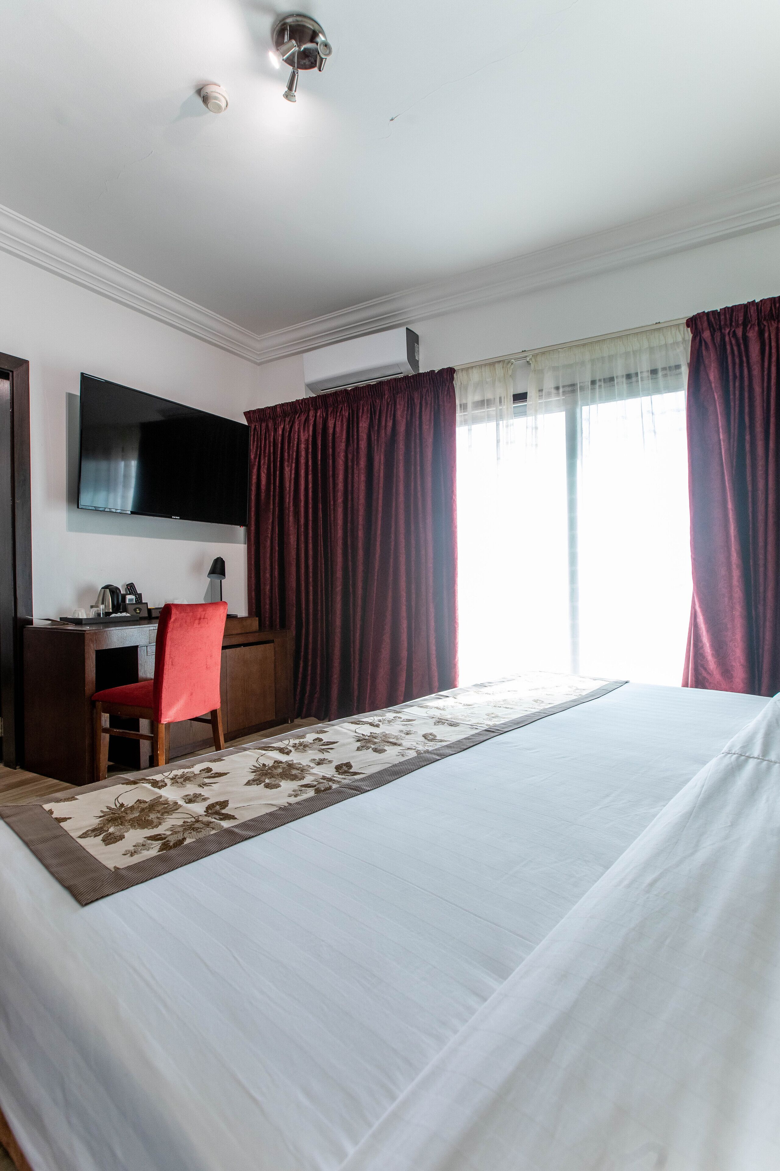A hotel room with a large bed, a desk with a chair, a flat-screen TV mounted on the wall, and a window with curtains.
