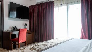 A hotel room with a large bed, a desk with a chair, a flat-screen TV mounted on the wall, and a window with curtains.