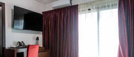 A hotel room with a large bed, a desk with a chair, a flat-screen TV mounted on the wall, and a window with curtains.