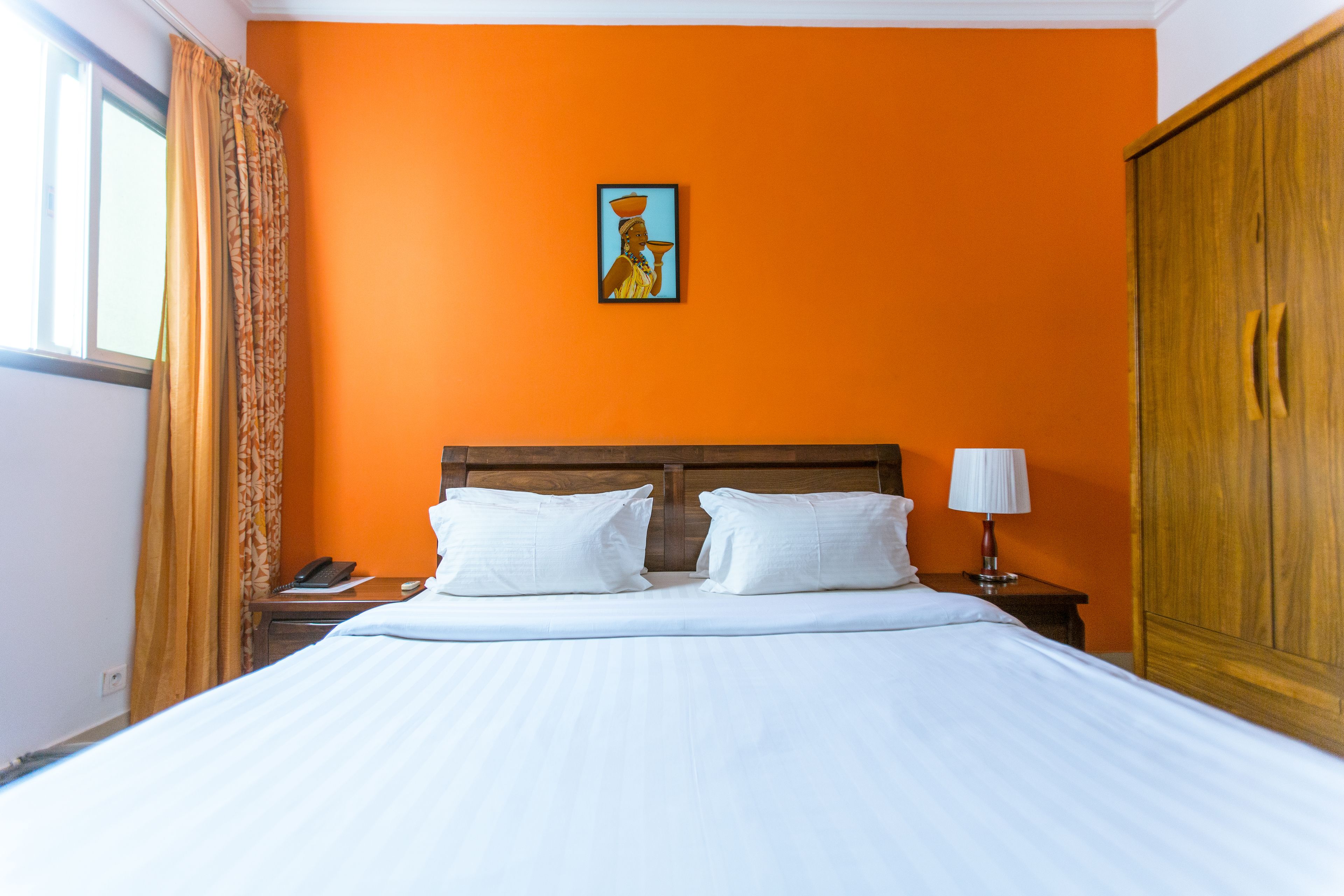 A hotel room with an orange wall, a wooden headboard, a bed with white linens, a nightstand with a lamp, and a painting on the wall.