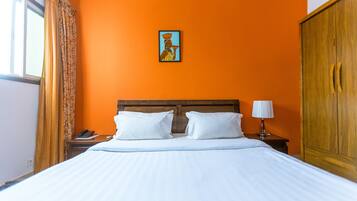 A hotel room with an orange wall, a wooden headboard, a bed with white linens, a nightstand with a lamp, and a painting on the wall.