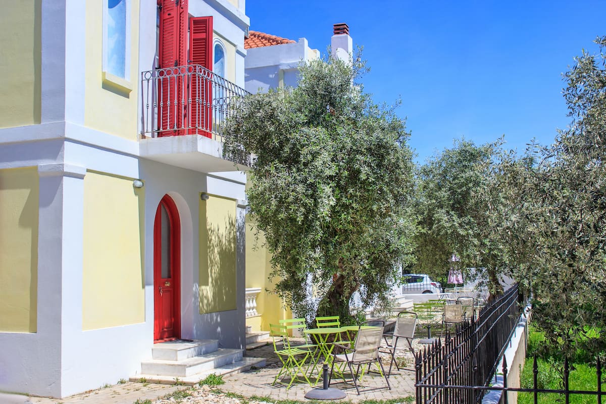 Un bâtiment avec une porte rouge et un balcon, des places assises à l’extérieur et un arbre.