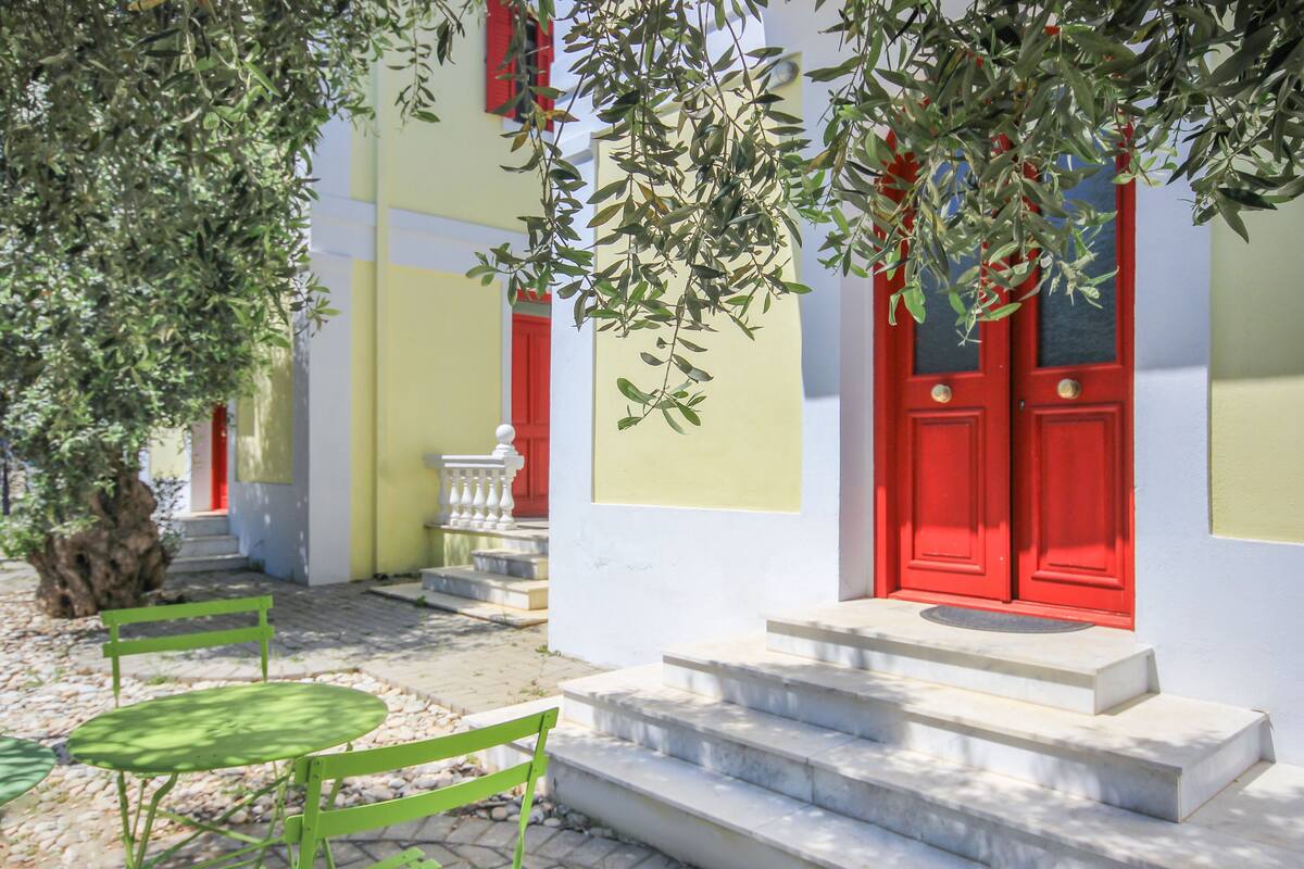 Un bâtiment avec une porte rouge, un escalier, et un ensemble de table et de chaises vertes placé à l’extérieur.