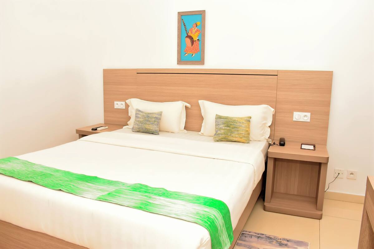 A neatly made bed with a green throw, a wooden headboard, and a small bedside table with a phone and a lamp.