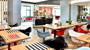 Un espace repas moderne avec des tables en bois et des chaises colorées, un coin salon avec des canapés et des fauteuils rouges, et une bibliothèque remplie de livres et de plantes.