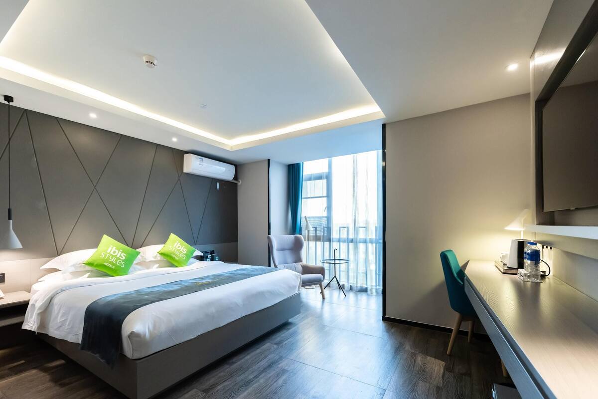 A modern hotel room with a large bed, a desk, a chair, and a view of the outdoors.