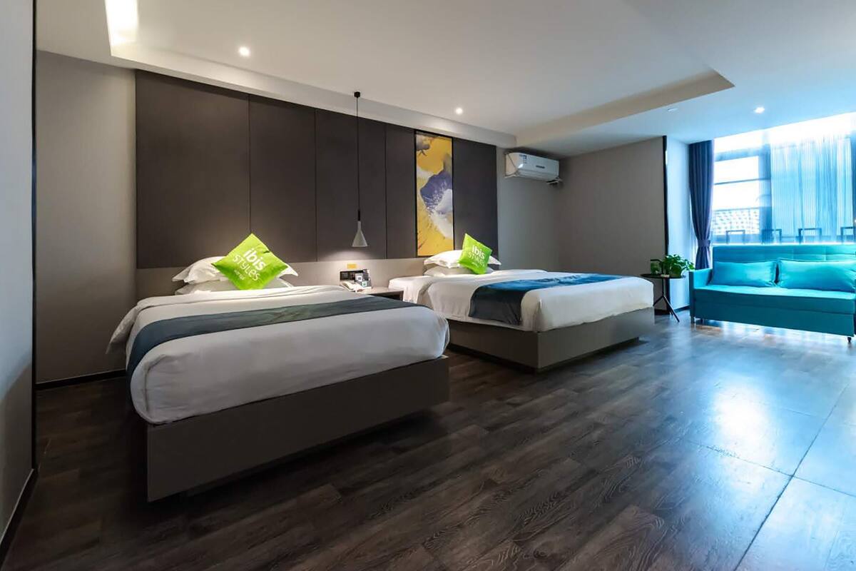 A modern hotel room with two beds, a sofa, and a large window.