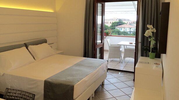 Habitación de hotel con cama, escritorio, silla, balcón con mesa y sillas, y vistas a edificios y árboles.