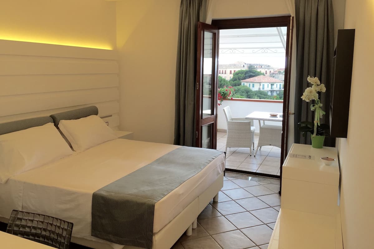 A hotel room with a bed, a desk, a chair, a balcony with a table and chairs, and a view of buildings and trees.