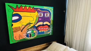 A room with a bed, a wooden headboard, a colorful abstract painting on the wall, and a window with curtains. - Jump in Bed Ipanema 10 - Loft (Rio de Janeiro)