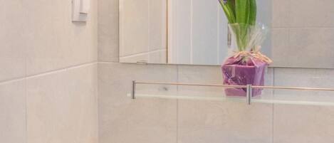 Un baño con un lavamanos blanco, un espejo y un florero con flores moradas sobre una repisa.