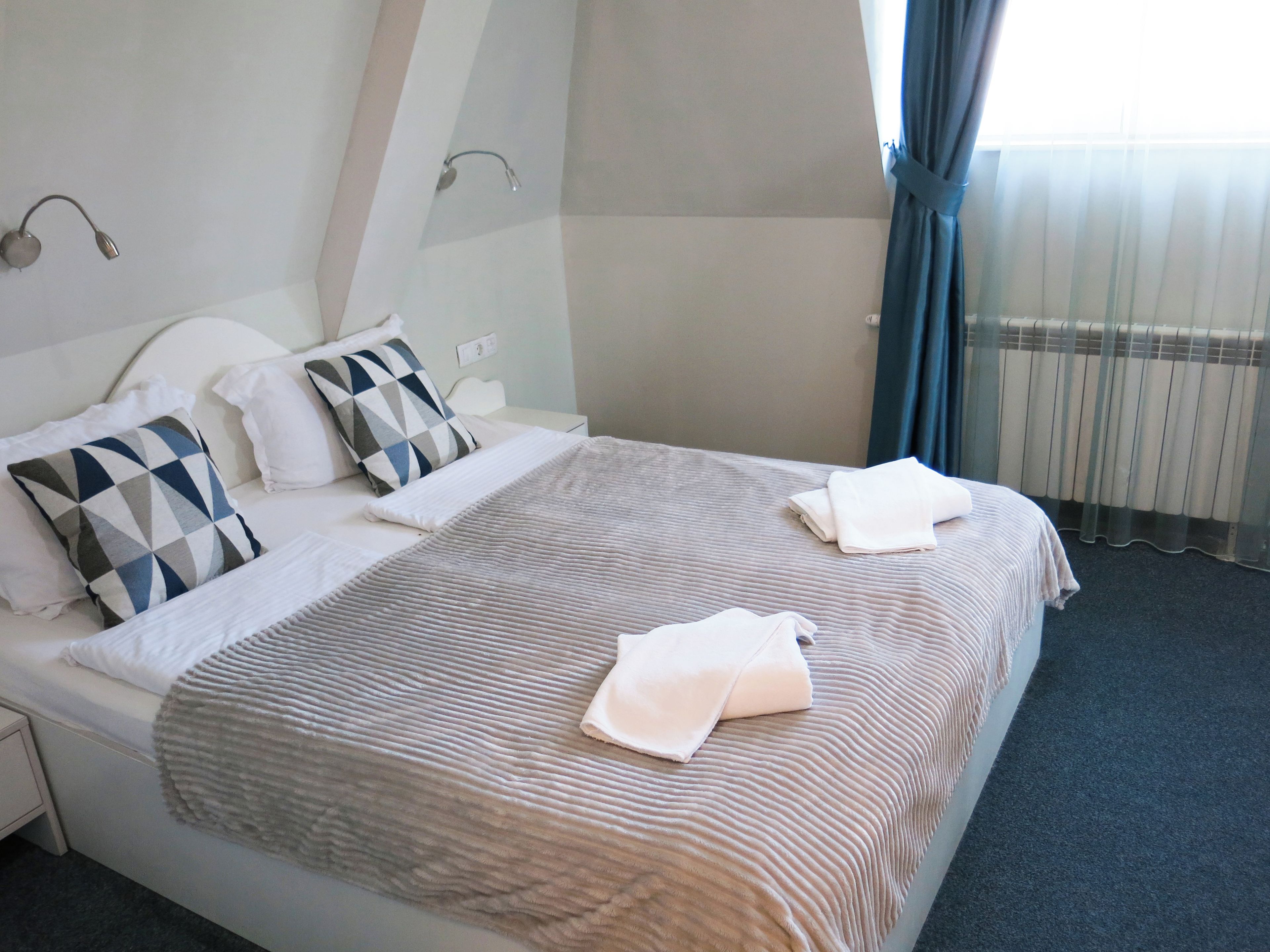 Habitación de hotel con cama, almohadas, manta, mesita de noche y ventana con cortinas.