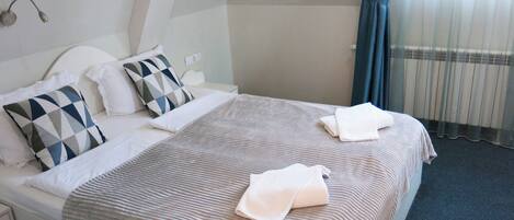 Habitación de hotel con cama, almohadas, manta, mesita de noche y ventana con cortinas.
