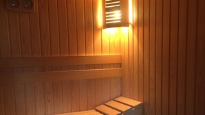 Una sauna de madera con bancos y suelo inclinado, un reloj en la pared y una lámpara.