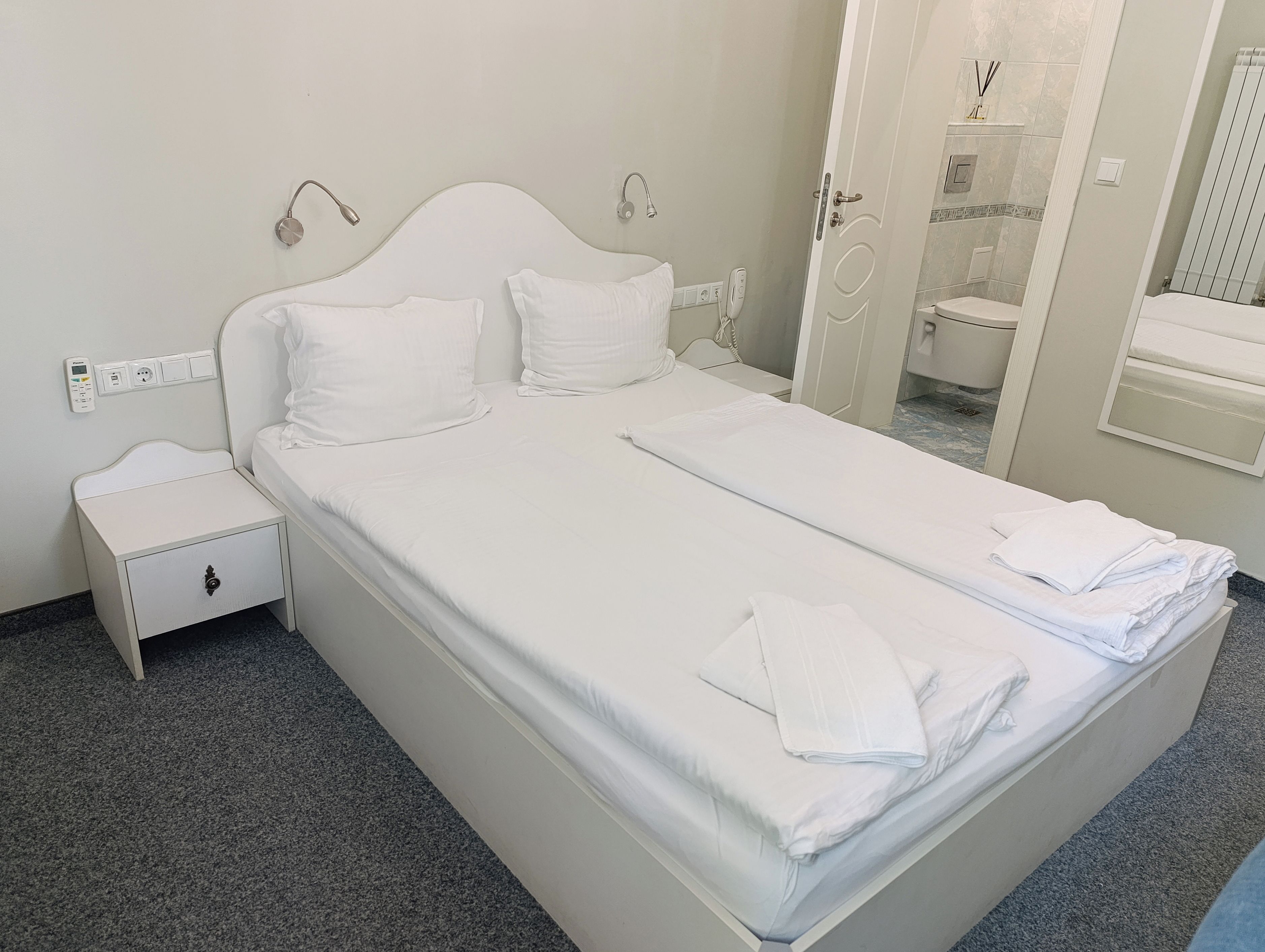 A hotel room with a bed, a nightstand, a bathroom with a toilet and sink, and a mirror.