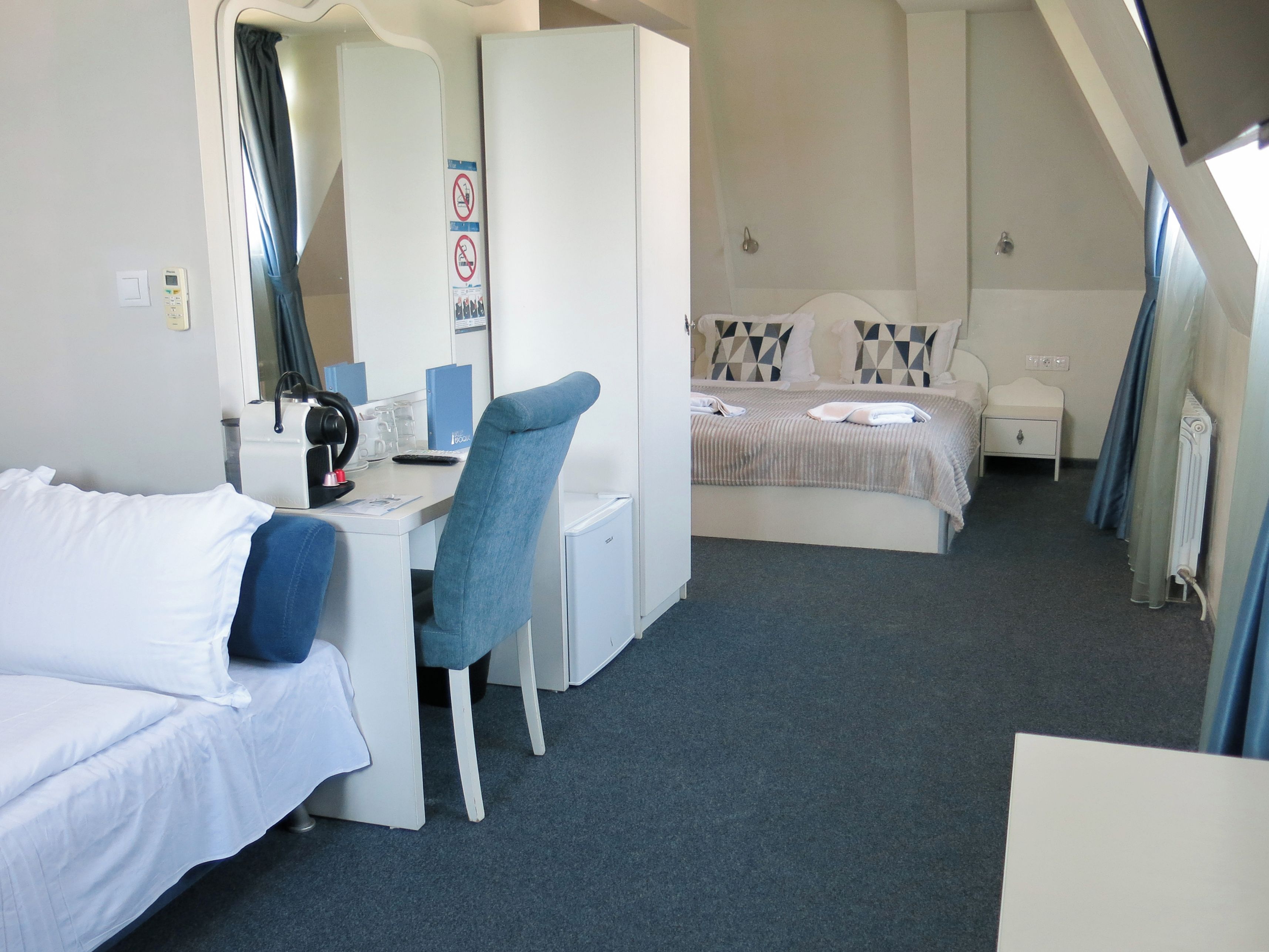 A hotel room with a bed, a desk, a chair, a small bedside table, and a radiator.