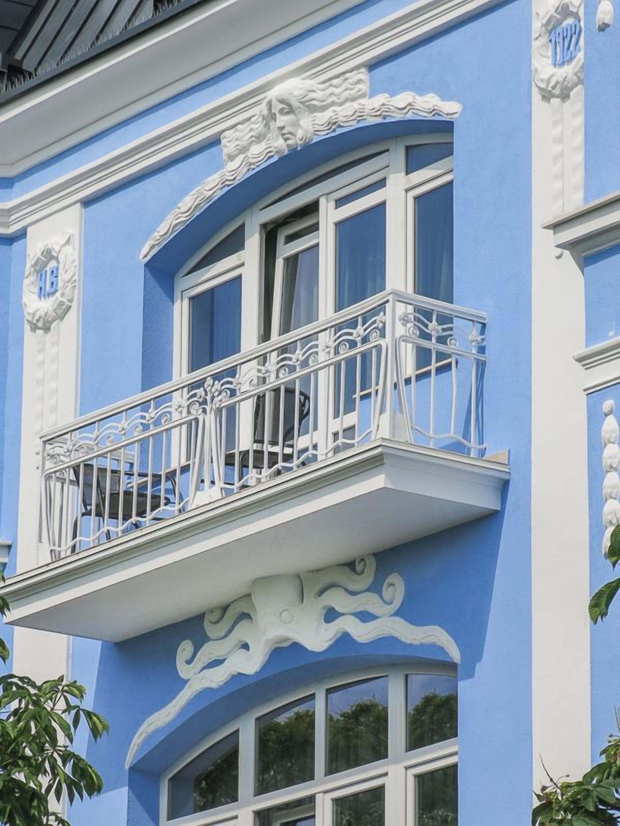 En bygning med en blå facade, en balkon med detaljerede hvide gelændere og dekorative hvide arkitektoniske detaljer.