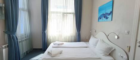 A hotel room with a double bed, a bedside table, a wall-mounted lamp, a painting, and a window with curtains.