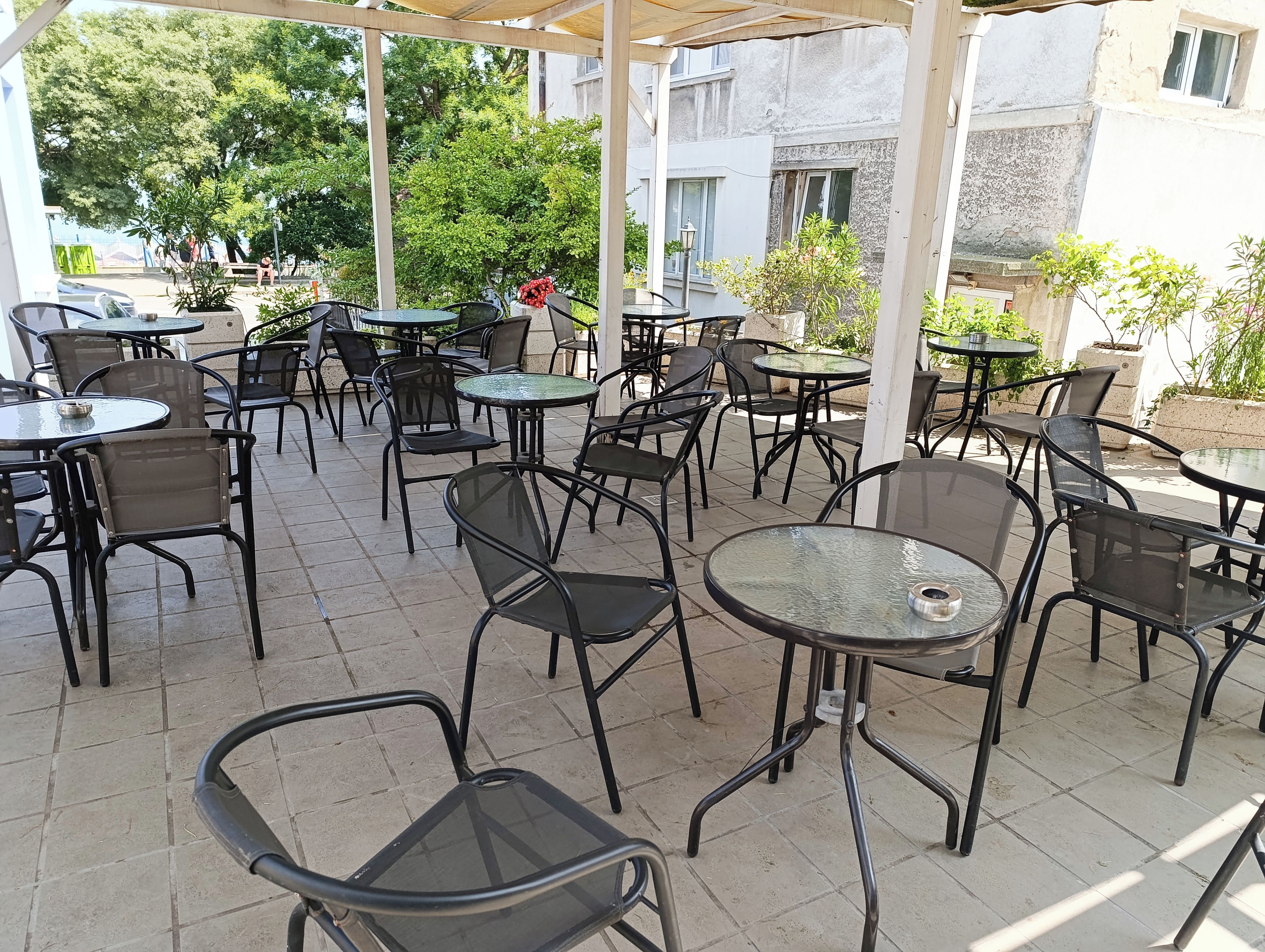 An outdoor café with multiple tables and chairs, surrounded by greenery and buildings.