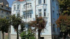 Un edificio azul claro con detalles arquitectónicos ornamentales, incluyendo un balcón y marcos de ventanas decorativos.