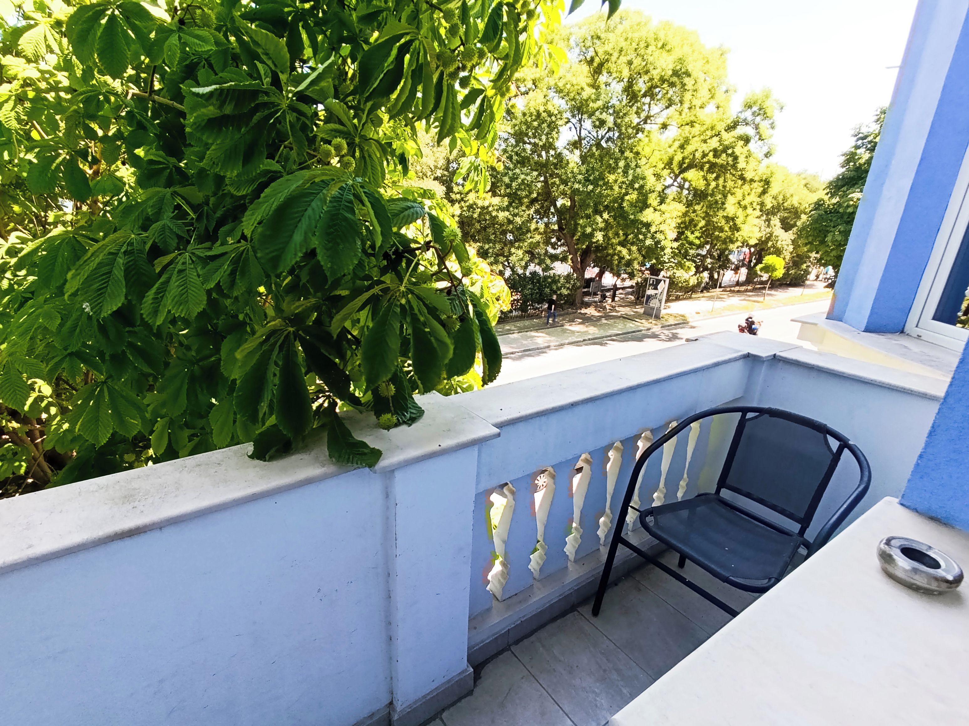Un balcón con una silla de metal, barandal blanco y vista a árboles y una calle.