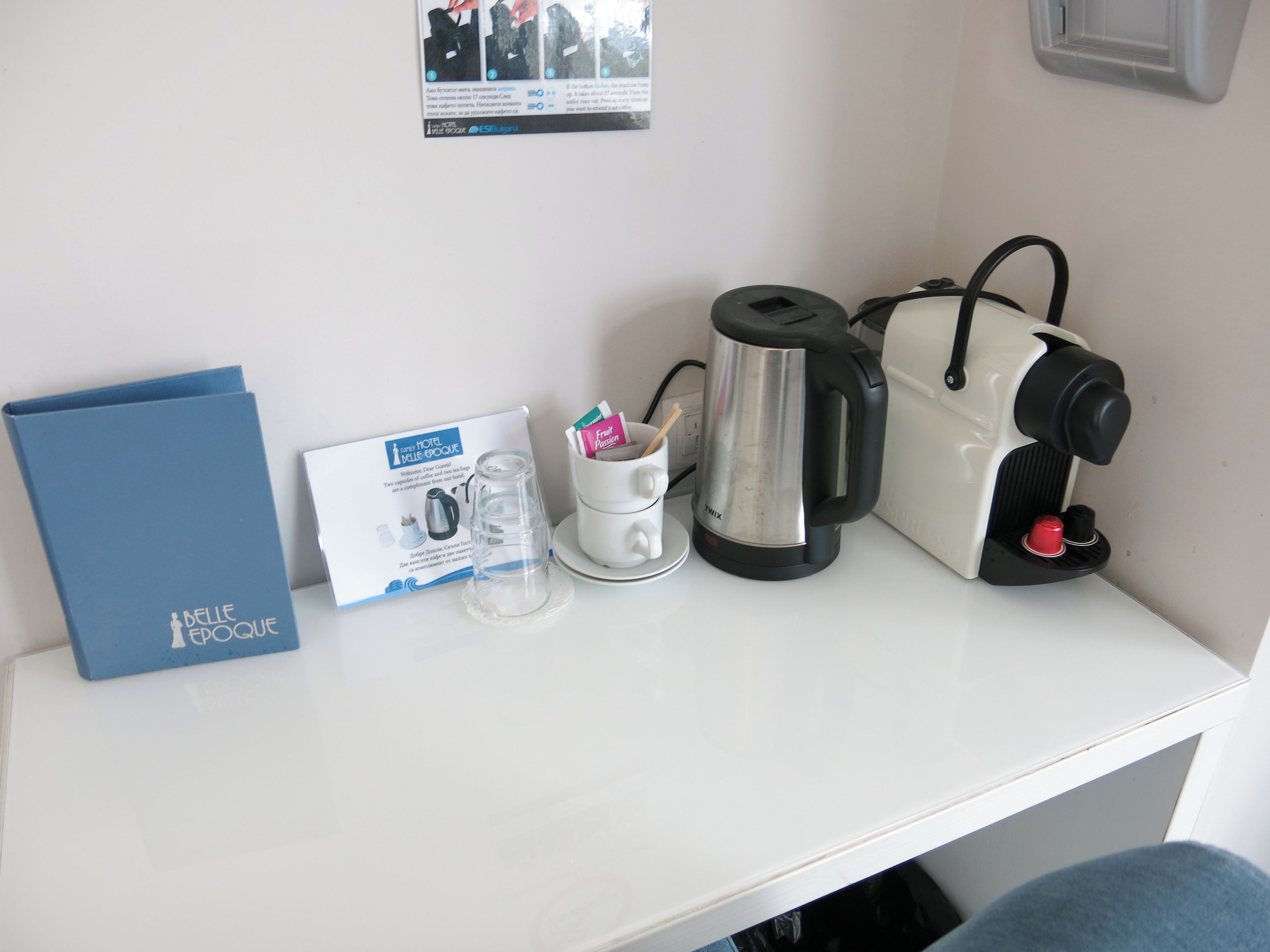 A white table with a blue booklet, a glass mug with a spoon, a stainless steel coffee pot, and a white electric kettle.
