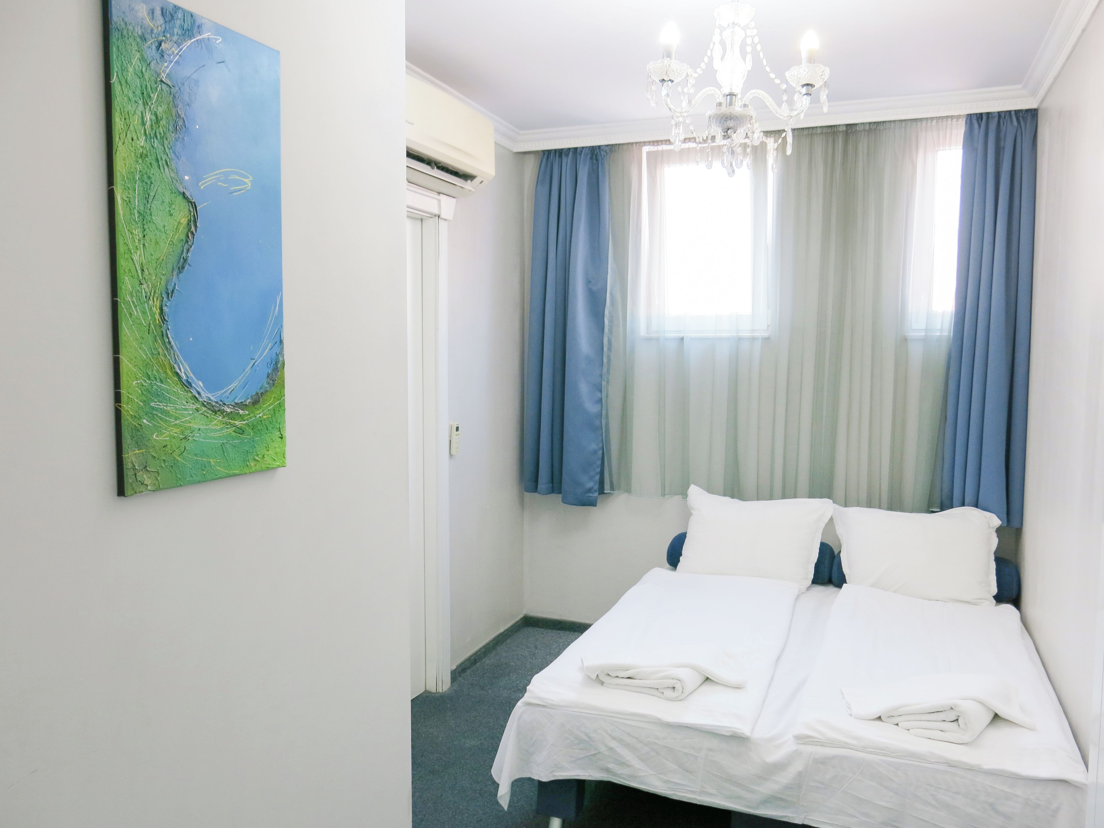 A hotel room with a bed, a chandelier, a painting, and a window with curtains.