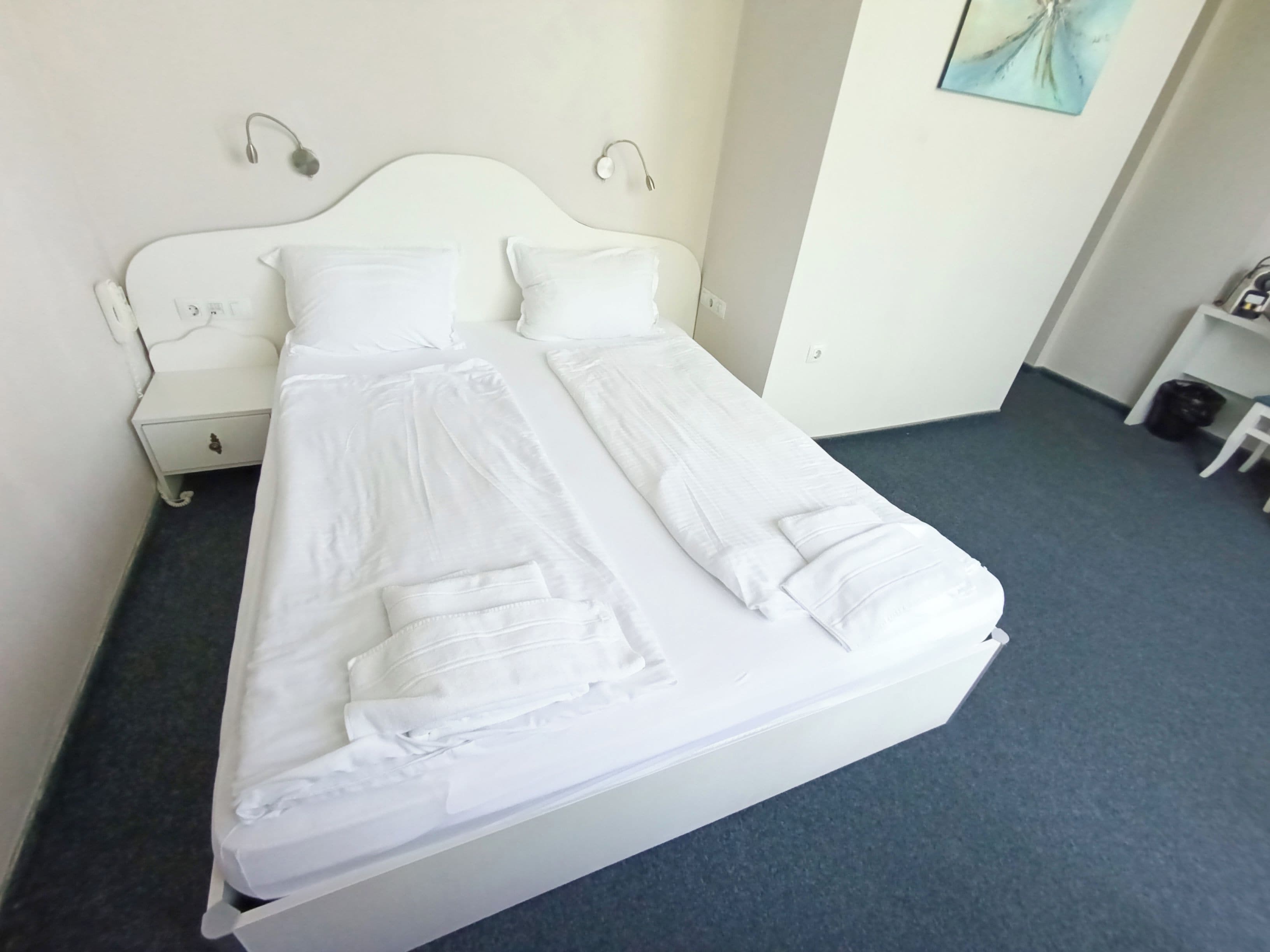 Un lit bien fait, avec du linge de lit blanc et des oreillers, une table de chevet blanche dotée d’un tiroir, et un luminaire fixé au mur.
